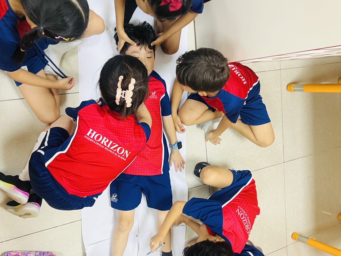 Seen from above, a group of students trace around a classmate lying on a long sheet of paper.