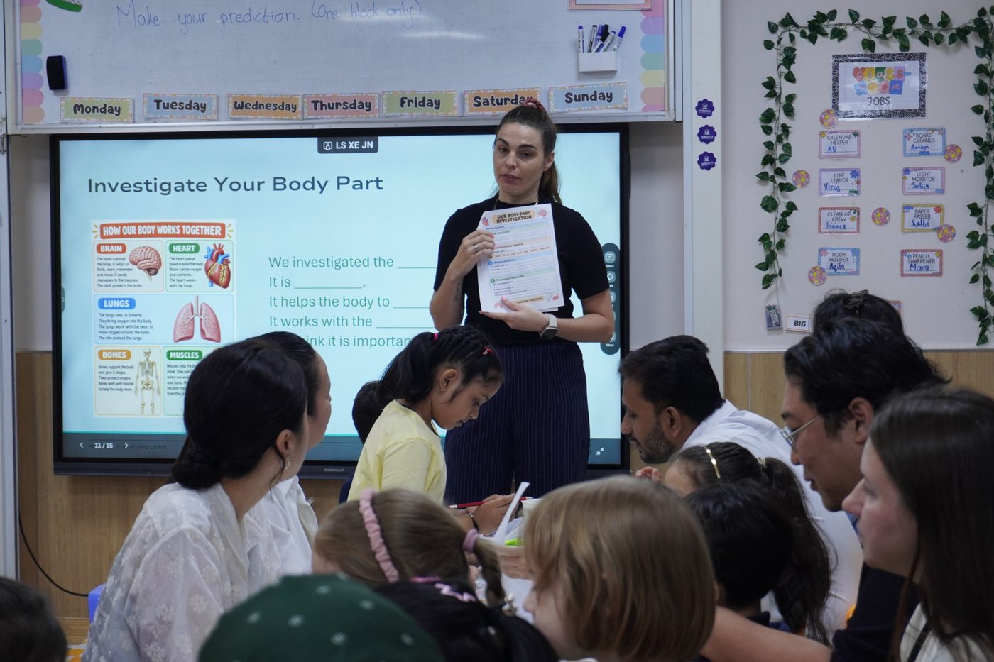 A teacher holds up an investigation worksheet in front of a screen while parents sit with their children at classroom tables.