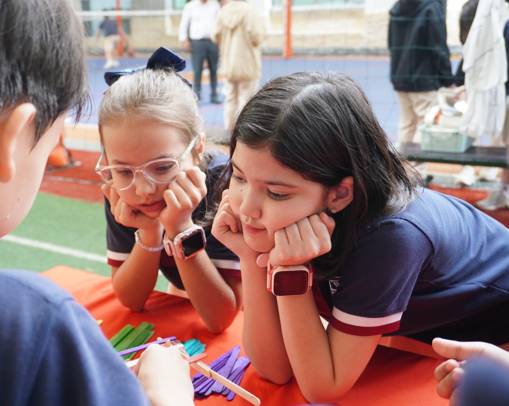 Two primary students rest their chins on their hands, watching closely as a classmate builds with coloured craft sticks.