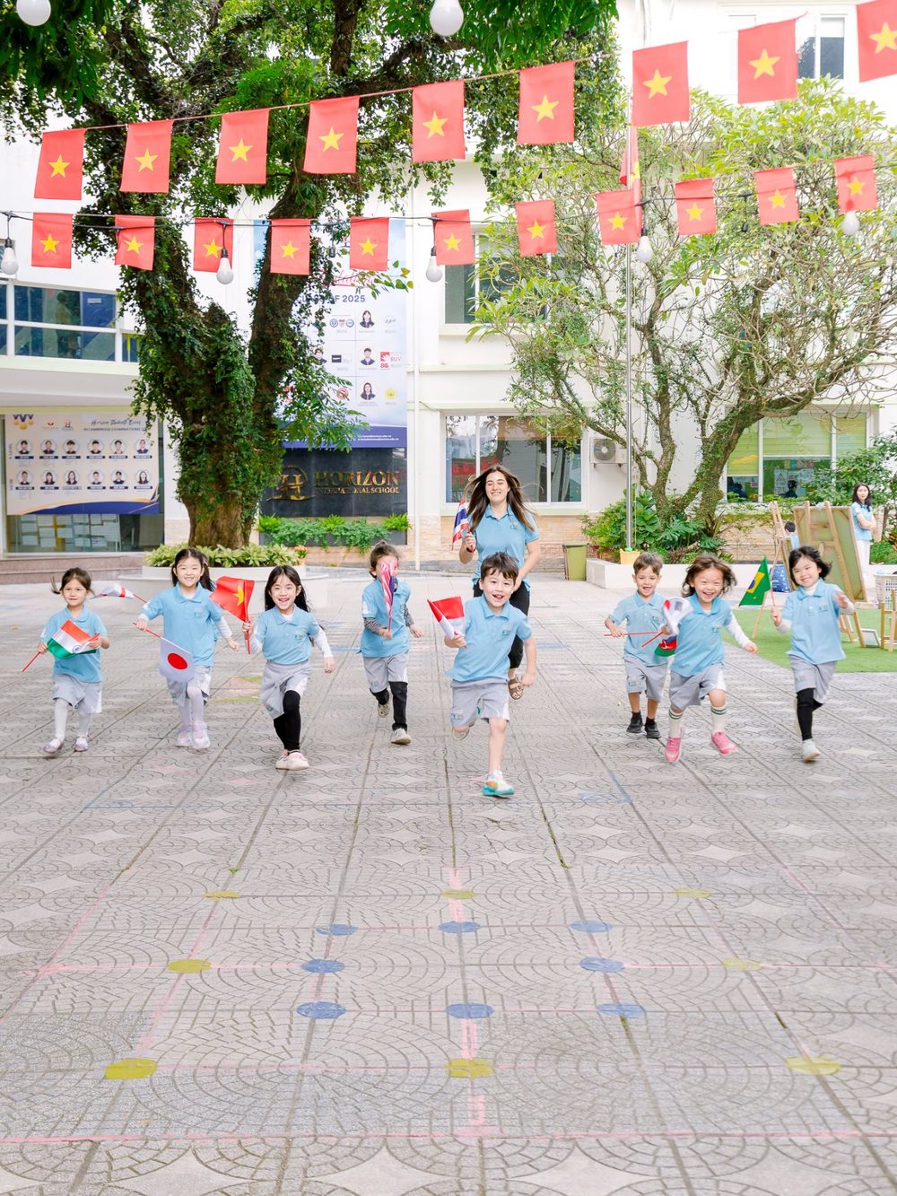 Kindergarten children run across the school courtyard holding flags of different countries, under strings of Vietnamese flags.