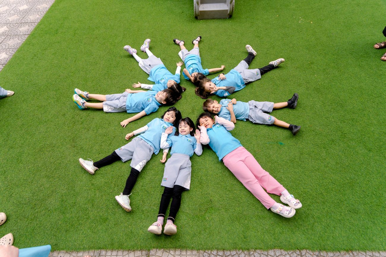 Kindergarten children lie on the grass with their heads together in a star shape.