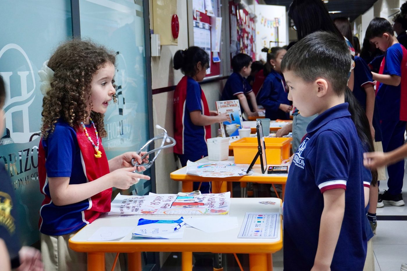 A student holding a stethoscope explains her poster to a classmate across a table.