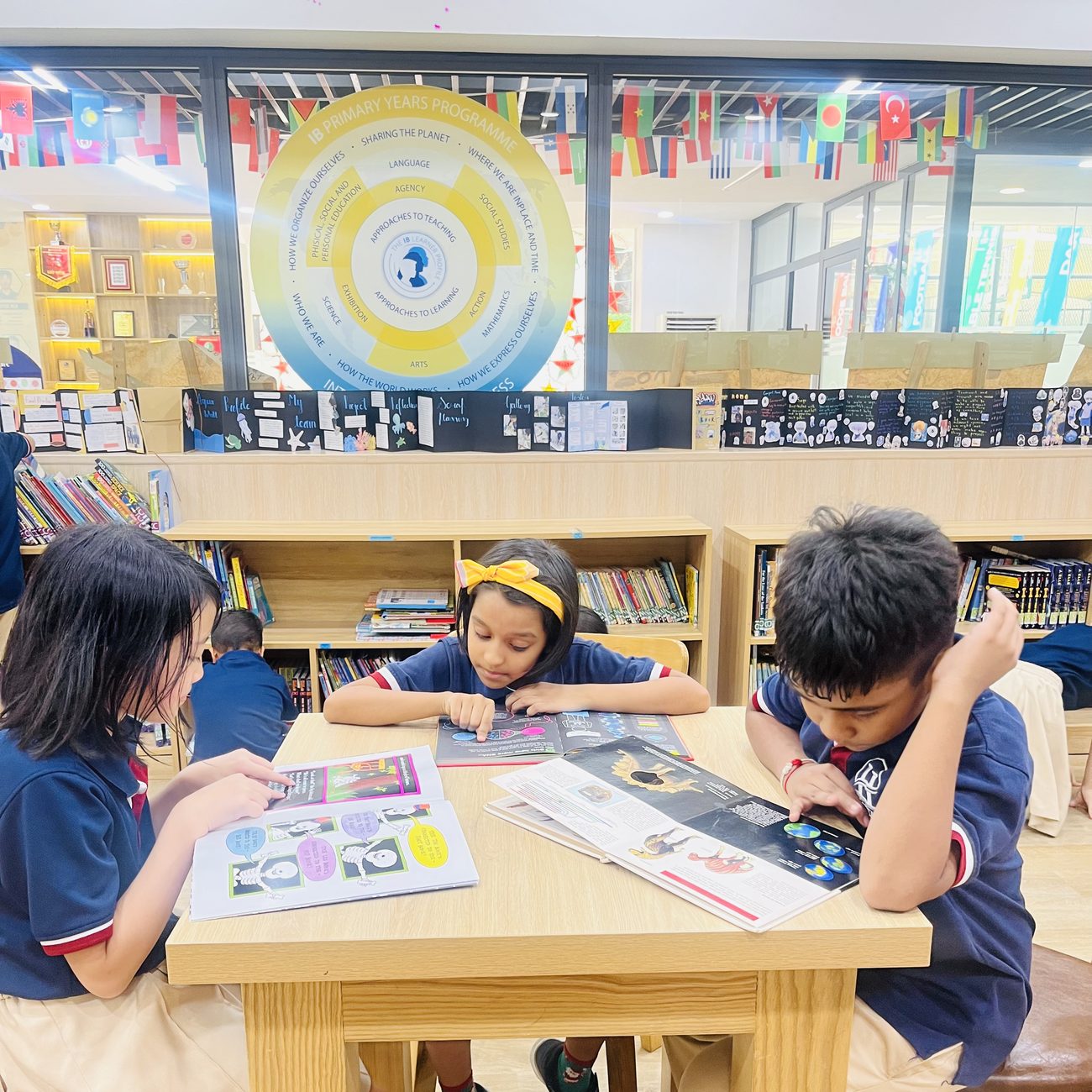 Three students read non-fiction books at a library table, beneath a large circular display of the IB Primary Years Programme framework.