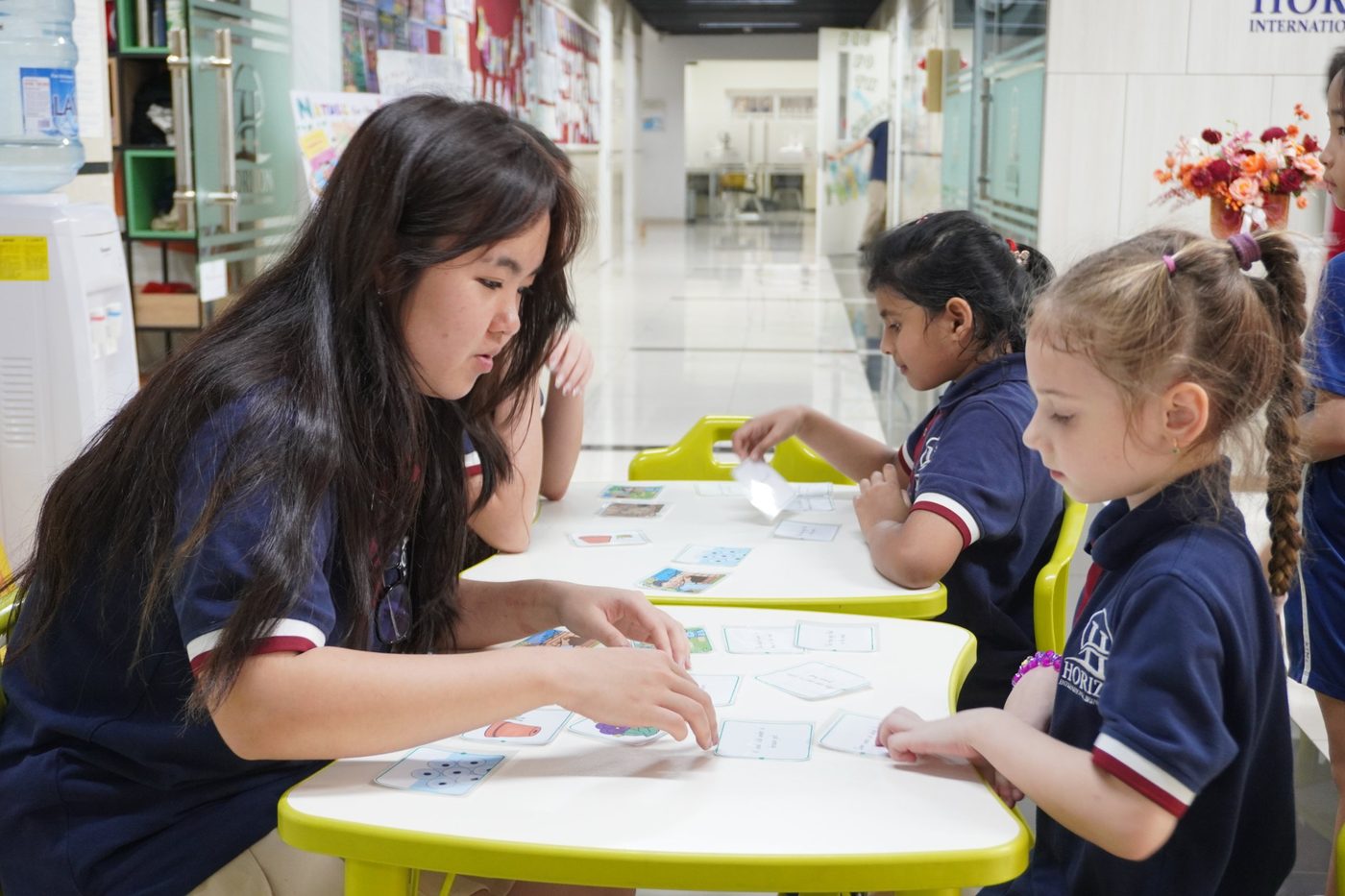 An older student lays out picture cards on a table for two younger students in the school corridor.