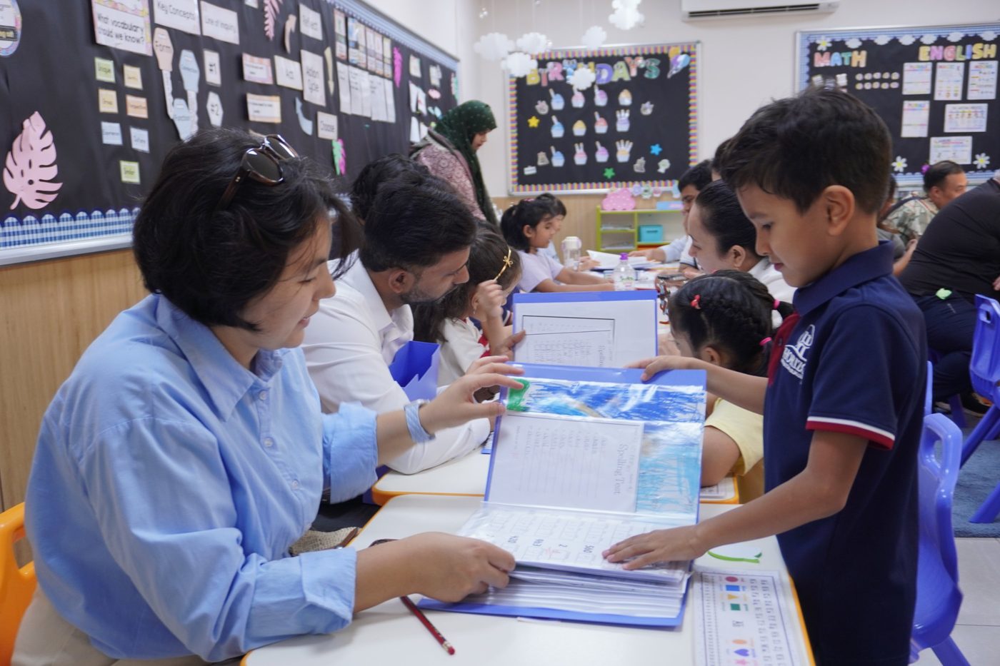 A student shows a parent the pages of his portfolio folder at a classroom table, while other families do the same behind them.