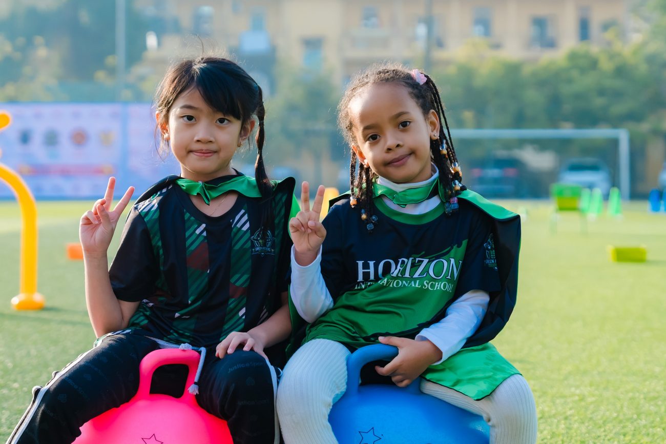 Two students in green house kit sit on space hoppers on the sports field.