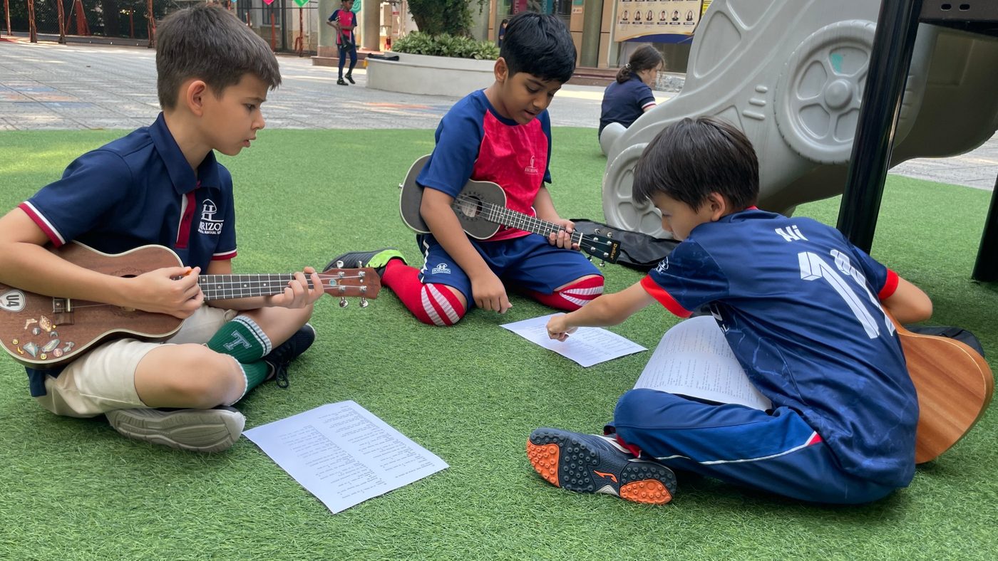 Three students sit on the grass practising ukulele from song sheets.