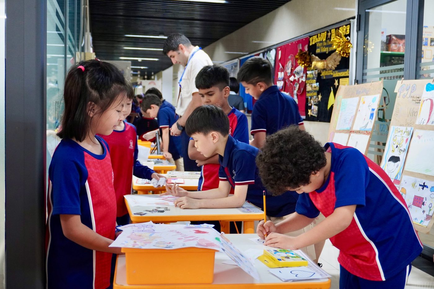 Students work on drawings at desks in the corridor, beside display boards headed PYP and Who we are.